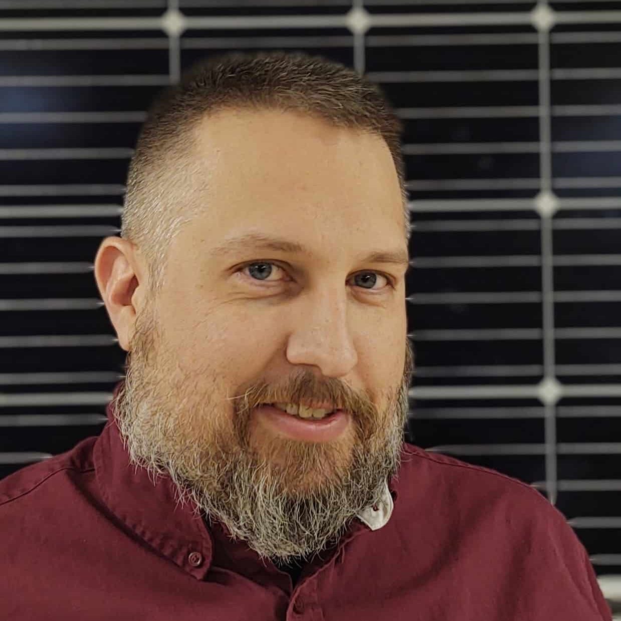  A smiling man with a neatly groomed beard and short hair, wearing a maroon shirt, in front of a geometrically patterned, black and white background.