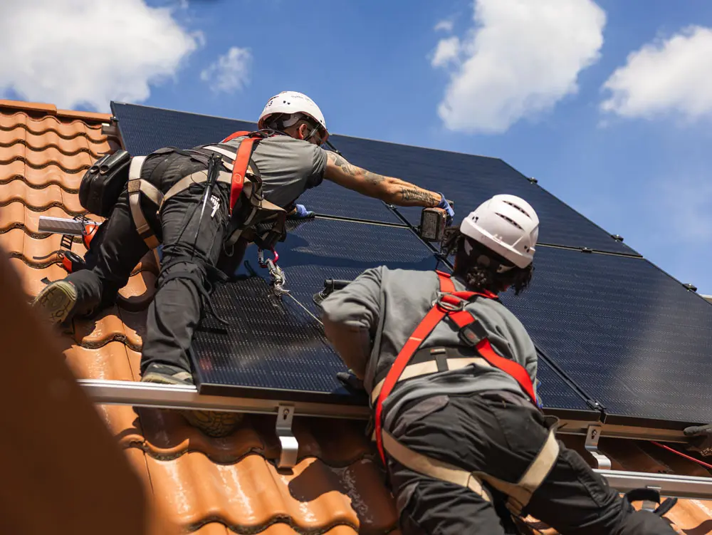 Zwei Infravoltaic Handwerker bei der Montage einer Solaranlage auf einem Hausdach mit hellbraunen Ziegeln