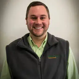  A smiling man in a green shirt with a black zip-up vest standing against a neutral background.