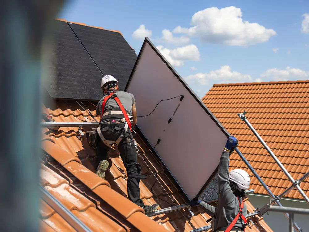 Zwei Infravoltaic Monteure bei der Montage einer PV-Anlage auf einem Hausdach mit hellbraunen Dachziegeln. Man sieht zwei fertig montierte Solarmodule und ein Solarmodul von der Rückseite, während es hochgetragen wird.