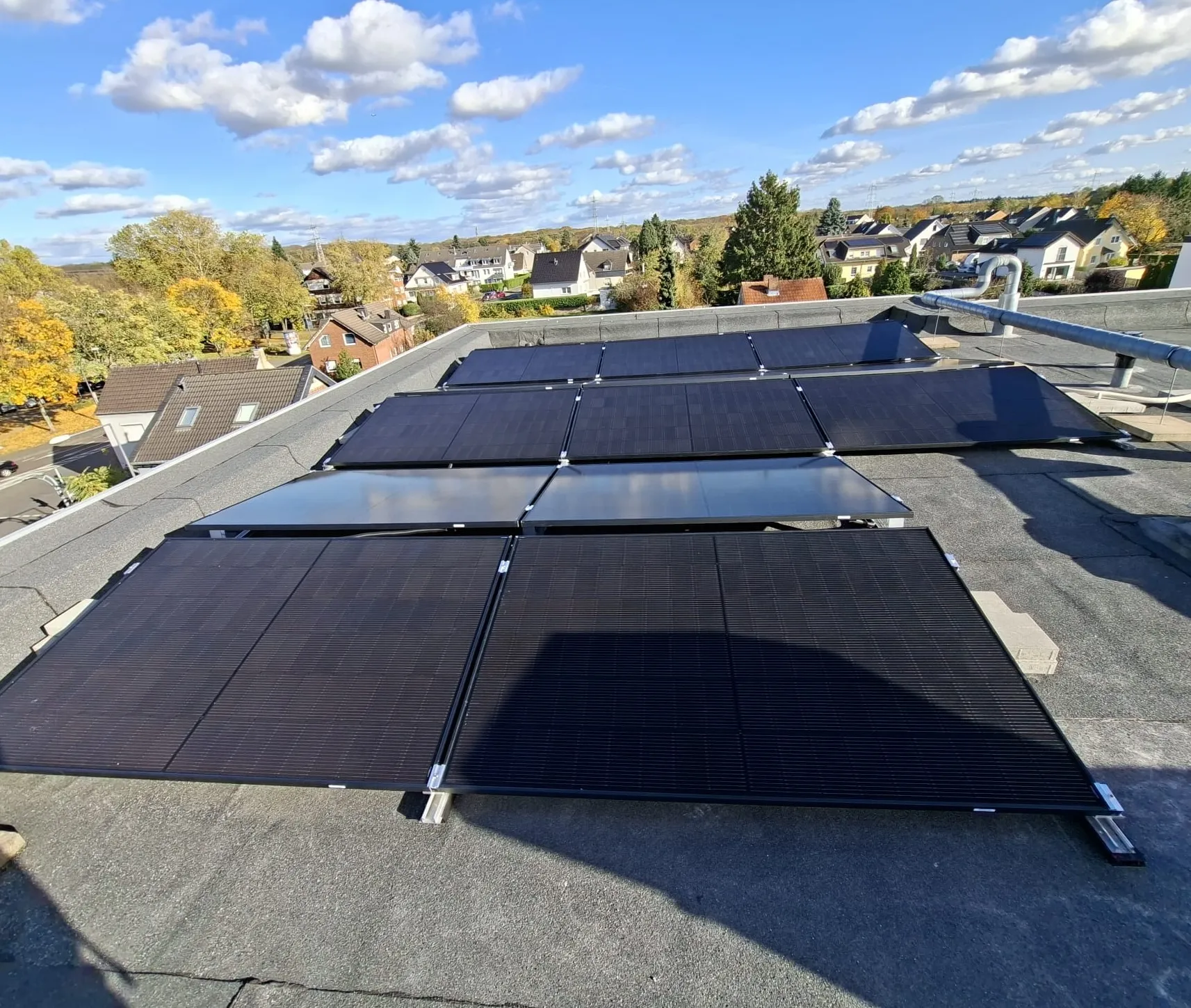 Solarmodule auf einem Flachdach bei sonnigem Himmel mit einer Wohnsiedlung und Herbstbäumen im Hintergrund.