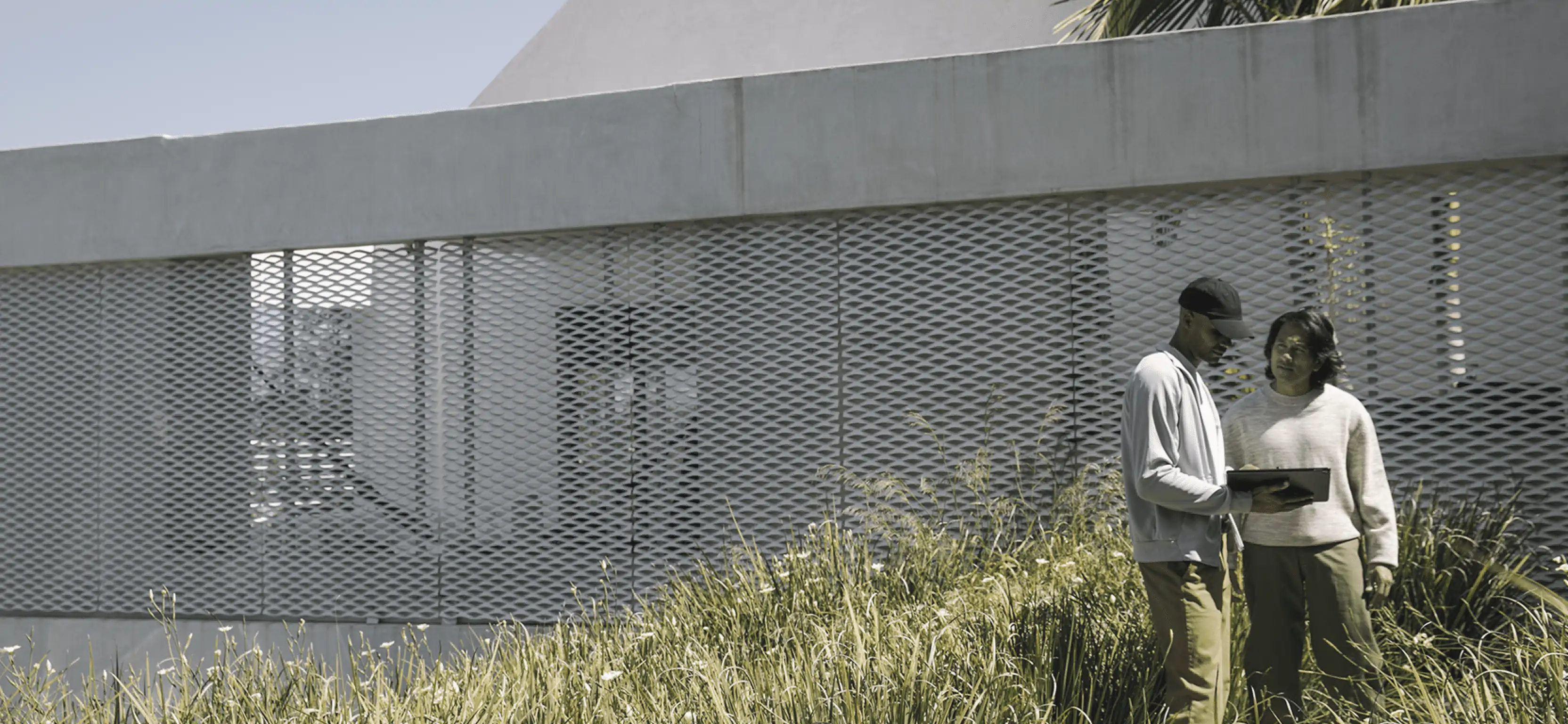 Two professionals having a discussion beside a modern building with a perforated metal facade, surrounded by tall grasses.