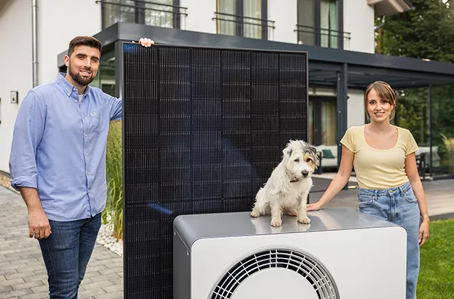 Infravoltaic Kundenfamilie mit Solarmodul und Wärmepumpe vor dem Haus. Auf der Wärmepumpe sitzt ein kleiner Hund