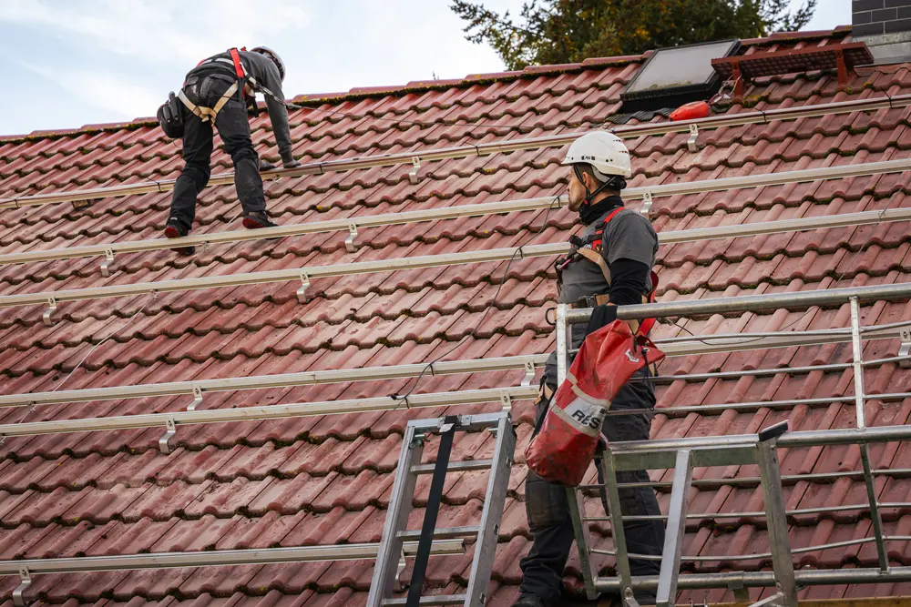 Zwei Infravoltaic Handwerker bei der Vorbereitung einer PV-Montage auf einem Hausdach mit hellbraunen Dachziegeln. Auf dem Dach sind schon Montageschienen für die Solarmodule befestigt.