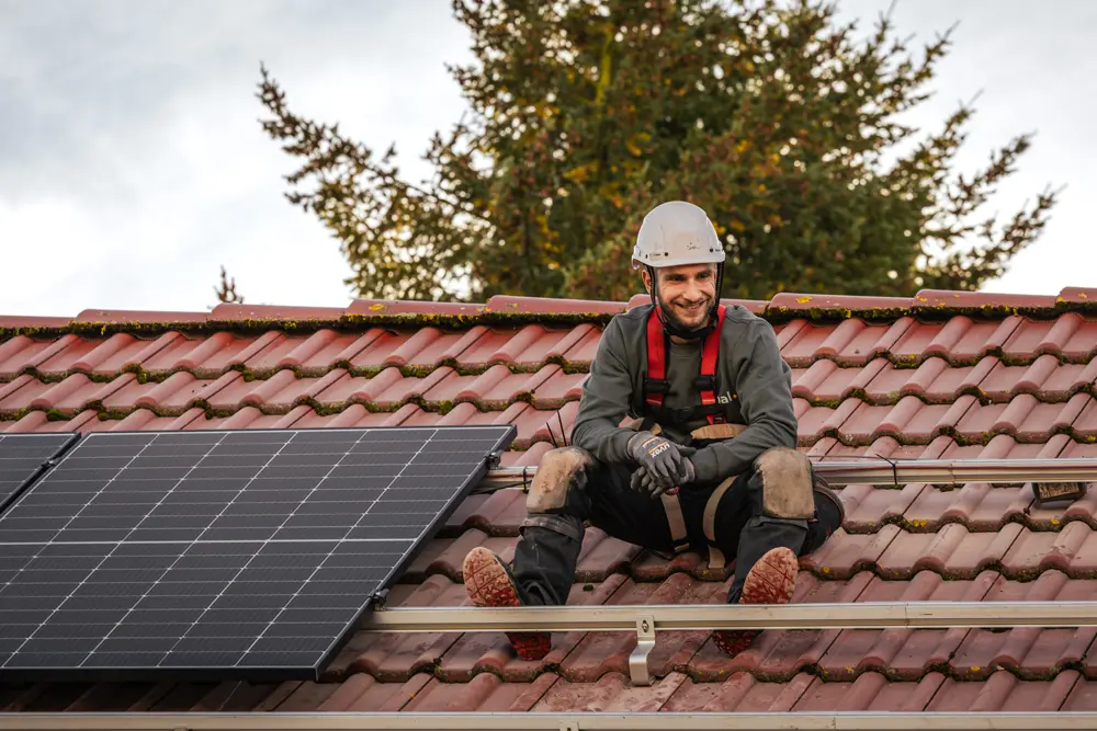 Infravoltaic Handwerker bei der Montage einer Solaranlage auf einem hellbraunen Dach. Im Hintergrund guckt ein Baum hinter dem Dach hervor