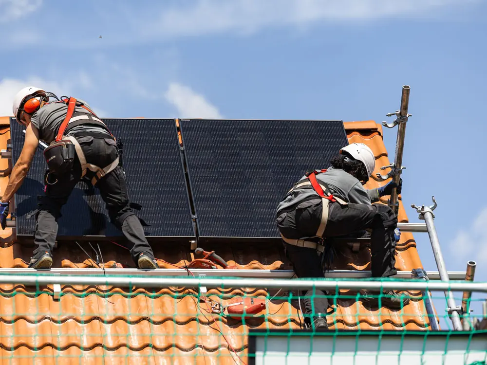 Zwei Infravoltaic Monteure bei der Montage einer PV-Anlage auf einem Hausdach mit hellbraunen Dachziegeln.