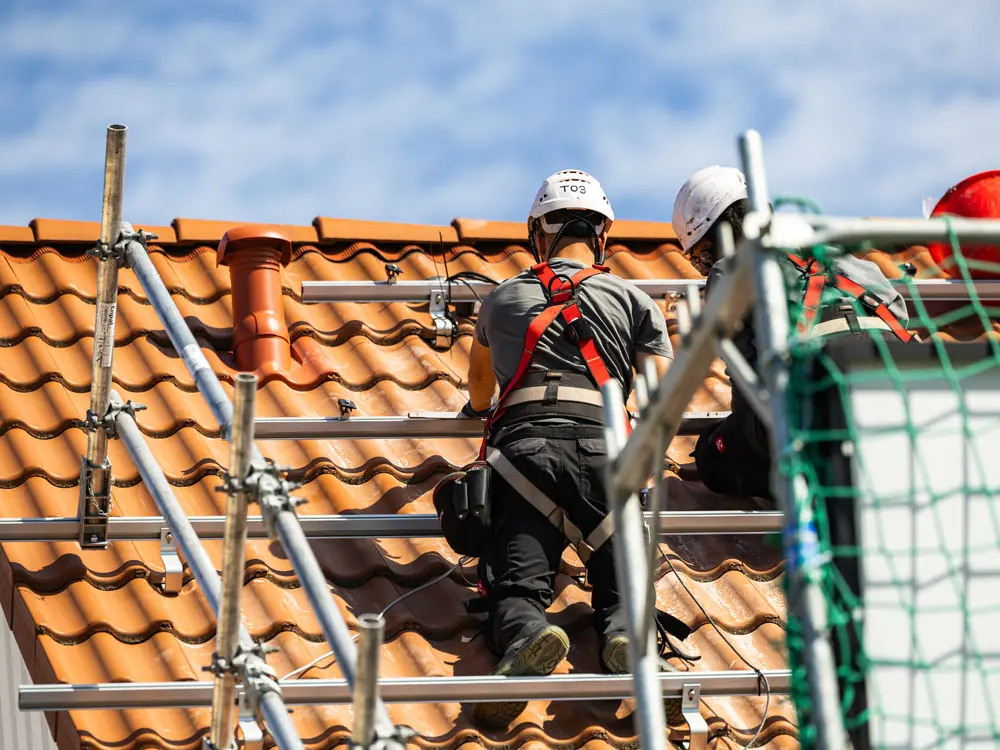 Zwei Infravoltaic Handwerker bei der Montage einer Solaranlage auf einem Hausdach mit hellroten Dachziegeln. Im Vordergrund sieht man den Fallschutz für die Handwerker mit einem grünen Netz.