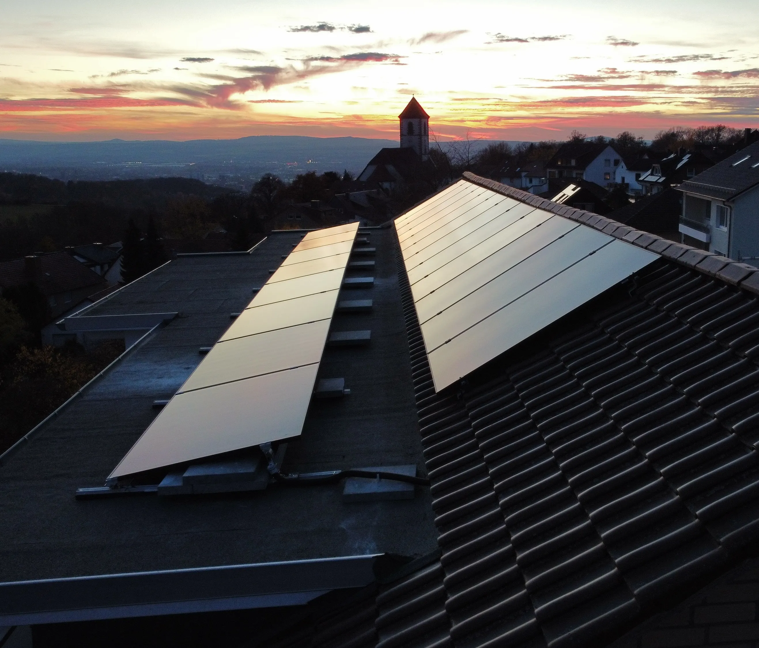Solarmodule auf zwei Hausdächern bei Sonnenuntergang mit einer Kirche im Hintergrund.